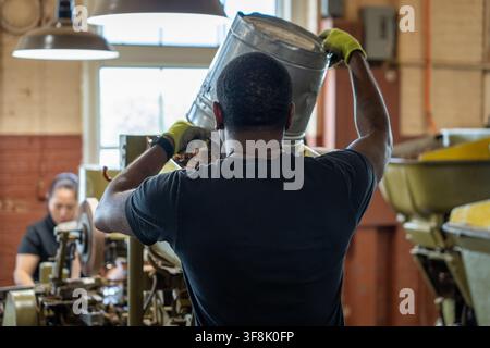 Tampa, FL, USA. 12th Aug 2026. A worker empties a metal pail of filler tobacco into the hopper of an antique 1930s cigar-making machine as an operator works at the next station in the machine-rolling area of J.C. Newman Cigar Co.'s El Reloj factory in Ybor City. Founded in 1895, fourth-generation J.C. Newman is America's oldest family-owned premium cigar maker, and the 1910 clock-tower building - part of a National Historic Landmark District - is the last operating historic cigar factory in the United States. Credit: Tosh Persons / Ģ����ý Live News Stock Photo