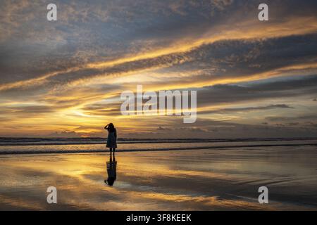 View of a woman standing on the wet sand of Inani Beach at sunset, with golden light reflecting on the water and a dramatic sky in Cox's Bazar Distric Stock Photo