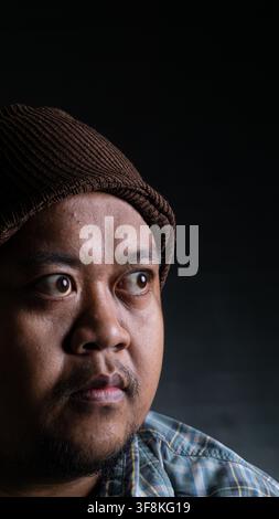 A close up portrait of a young Asian male from Indonesia Southeast Asia looking away to the side against a dark background, The thoughtful man wears a Stock Photo