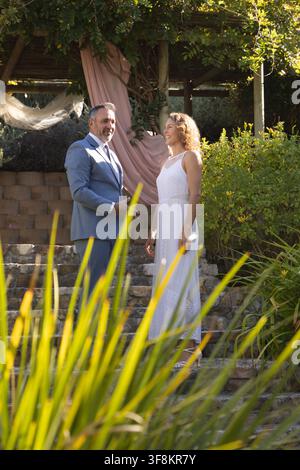 Couple standing on steps under pergola in garden, wearing suit and white dress with pink drapery Stock Photo