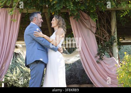 Couple embracing under pergola with pink drape and vines, wearing blue suit and white dress Stock Photo