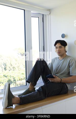 Adult Asian man sitting on window bench by sliding window holding phone wearing headset, copy space Stock Photo