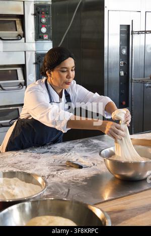 Female chef in coat and apron stretching dough over mixing bowl at kitchen workbench with flour Stock Photo