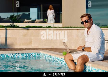 A relaxed man in white sits with his laptop at the edge of a private pool at a modern villa, sipping a green drink and blending remote work with leisure on a sunny summer day. Stock Photo