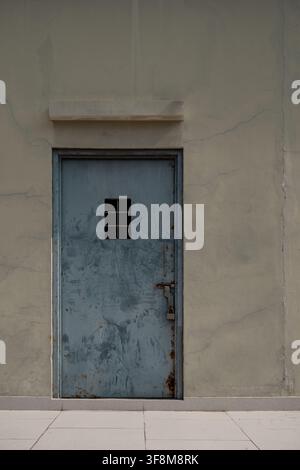 Rusty locked metal door with small window in cracked cement wall. Stock Photo