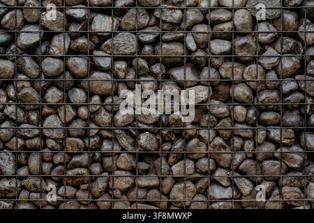 Dark grey river stones enclosed in a black wire mesh forming a gabion wall texture. Stock Photo