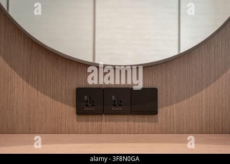 Black universal electrical power sockets and switches on wooden wall panel below round mirror. Stock Photo