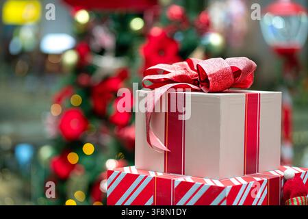 Christmas present in a box wrapped and tied with red ribbon. Christmas tree with decoration at the background. Stock Photo