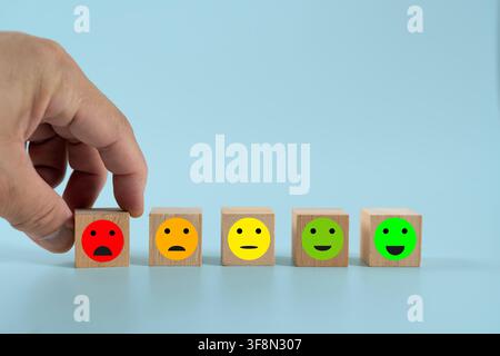 Hand pointing to the lowest satisfaction icon on wooden cubes, illustrating negative customer feedback, poor service rating, complaint handling, user Stock Photo