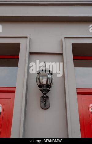 Classic black wall lantern mounted on a grey building exterior between red doors. Stock Photo