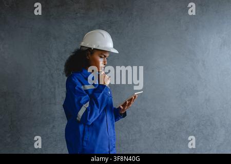 Portrait engineer woman wearing safety helmet and uniform standing gray background appears focused and professional representing modern technology con Stock Photo