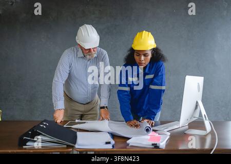 Engineer senior and woman engineer wearing safety helmets discussing blueprints in office collaborating on construction representing teamwork and prof Stock Photo