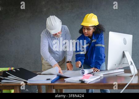 Engineer senior and woman engineer wearing safety helmets discussing blueprints in office collaborating on construction representing teamwork and prof Stock Photo