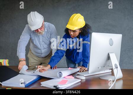 Engineer senior and woman engineer wearing safety helmets discussing blueprints in office collaborating. Stock Photo