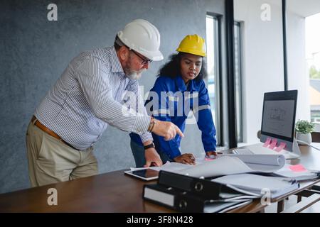 Engineer senior and woman engineer wearing safety helmets discussing blueprints in office collaborating. Stock Photo