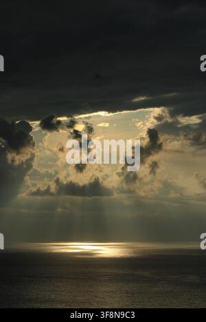Vertical seascape showing sunlight beams piercing dark storm clouds and reflecting on the ocean surface during sunset. Stock Photo