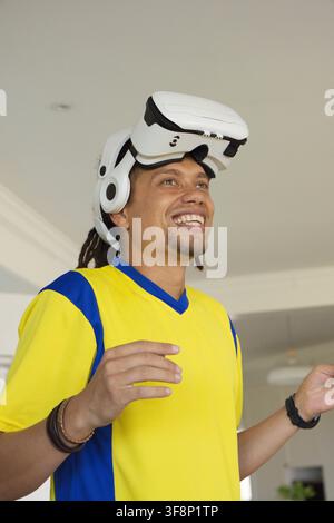 Mid adult African man wearing yellow blue jersey raising hands in living room with VR headset Stock Photo