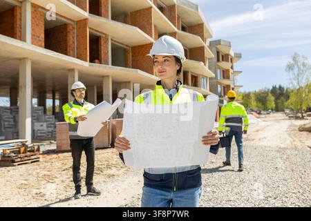 Confident female architect holding blueprint looking away with team Stock Photo