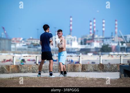 Matosinhos, Portugal - 29 July 2026: View of two young men practicing boxing with gloves on a promenade near the beach with industrial chimneys in the Stock Photo