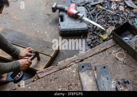 A carpenter hammering nail into wooden plank on workshop floor, rustic tools and saw blade scattered, focused hands working with drill case and hardwa Stock Photo