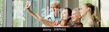 Diverse group of Caucasian senior and young women with senior man taking selfie on smartphone in gym. Image represents active lifestyle, suitable for senior fitness marketing Stock Photo