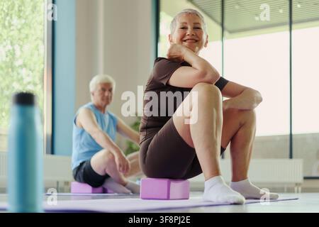 Portrait of smiling senior Caucasian woman sitting on yoga block during fitness class with senior man in background. Image represents active aging and wellness marketing campaigns Stock Photo