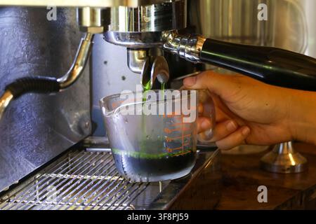 A green espresso shot extracted into glass measuring cup from portafilter, closeup of barista hand and metal espresso machine with crema and vivid gre Stock Photo