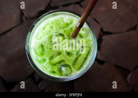 An iced matcha latte with straw and ice cubes in glass on wooden table, refreshing green beverage with froth and condensation Stock Photo