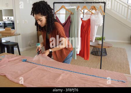 Adult woman cutting pink sheer fabric on large table with scissors, measuring tape in home studio. Sewing, tailoring, garment, textile, atelier, rack, Stock Photo