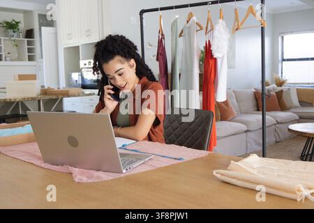Adult woman sitting at home workspace, holding smartphone, working on laptop with measuring tape Stock Photo