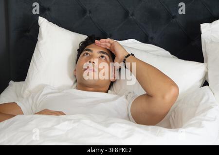 Asian man lying on back wearing white T-shirt and black beaded bracelet, looking upward on bed Stock Photo