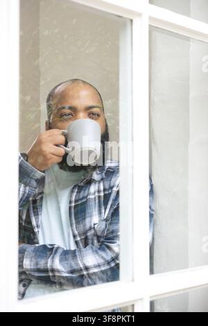 Bearded man in plaid shirt holding ribbed grey mug, sipping, looking through white window at home Stock Photo