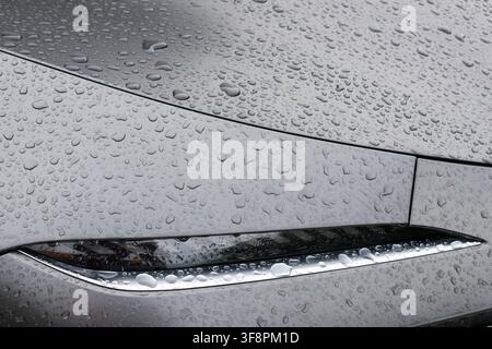 A sleek silver car body with water droplets on hood and aerodynamic headlight detail, moody rainy texture and reflective metallic surface Stock Photo