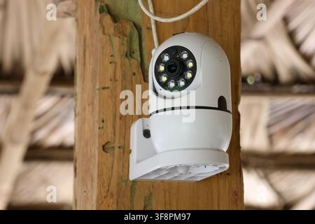 A white dome security camera mounted on wooden post with cable and night lights, surveillance device watching outdoor area with rustic thatch roof and Stock Photo