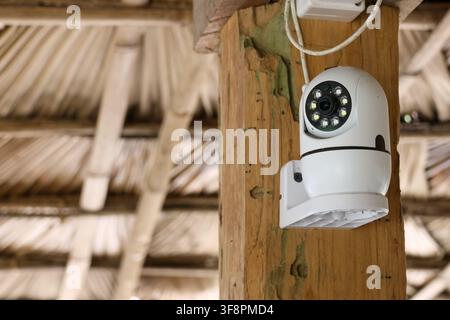 A white surveillance camera mounted on wooden post under thatched roof, security device watching outdoor area with visible wiring and infrared leds Stock Photo