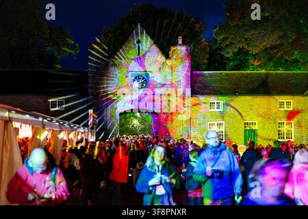 FESTIVAL CROWD AND A PSYCHEDELIC LIGHT SHOW, REPORTAGE, GREEN MAN FESTIVAL 2016, BRECON, WALES, UK, 2016: At night, a crowd of festival-goers walks past vendor tents towards an old stone building which is lit up with a vibrant, multi-coloured sunburst light projection. Photo: Rob Watkins. INFO: The Green Man Festival began in 2003 in Wales. Held annually in the Brecon Beacons on August’s third weekend, it celebrates alternative music, arts, literature, science and family-friendly culture in a beautiful mountain setting. Stock Photo