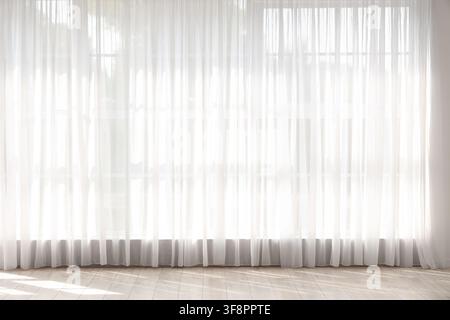 White sheer curtains covering a large window with bright sunlight streaming through onto a light wooden floor, the light creates soft shadows and high Stock Photo