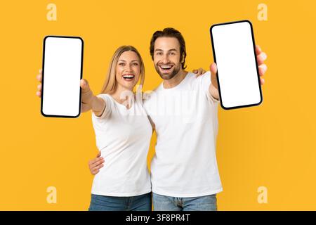 Happy Young Couple Holding Smartphones With Blank Screens, Yellow Background Stock Photo
