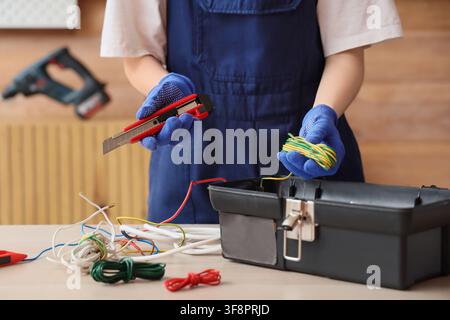 Person in blue work gloves holding a utility knife and electrical wires over a toolbox on a wooden table, a black toolbox and various colored wires ar Stock Photo