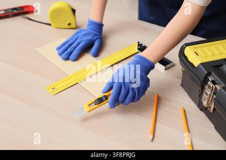 Person in blue gloves cutting wood veneer with a utility knife and ruler on a light wooden workbench Stock Photo
