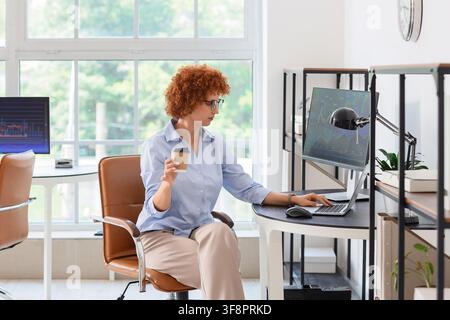 Young businesswoman with curly red hair analyzing financial data on a laptop and desktop monitor in a modern office, She holds a takeaway coffee cup, Stock Photo