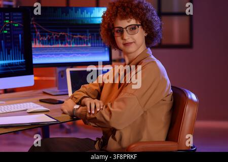 Female stock market trader with curly red hair and glasses working at night with multiple screens displaying financial data Stock Photo