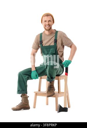 Smiling young lumberjack or gardener in green overalls and work gloves sitting on a wooden stool with an axe isolated on white background Stock Photo