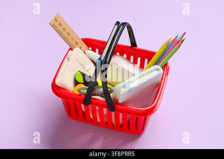 Red shopping basket filled with school supplies including pencils, a ruler, and notebooks on a purple background Stock Photo