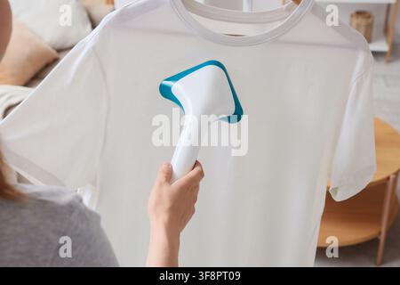 Person using a handheld garment steamer to remove wrinkles from a white t-shirt on a hanger laundry and clothing care, this image modern laundry solut Stock Photo