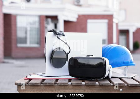 Construction site safety equipment with virtual reality headset and laptop on a wooden bench outdoors, this setup represents modern technology integra Stock Photo