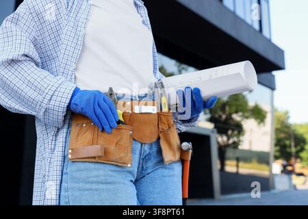Construction worker wearing blue gloves and a tool belt holding rolled blueprints at a building site Stock Photo