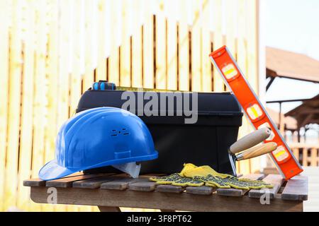 Blue hard hat, black toolbox, and construction tools on a wooden bench with a light wooden fence background Stock Photo