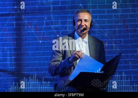 Smiling male speaker wearing a headset and holding documents in front of a blue brick wall with data projections Stock Photo