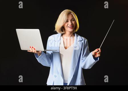 Smiling blonde woman in a light blue blazer holding a laptop and a pointer stick, presenting a business Stock Photo